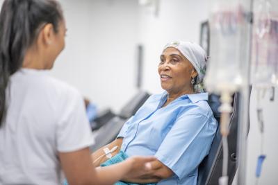 An image of a patient in a hospital gown sitting on a chair chatting with a nurse.